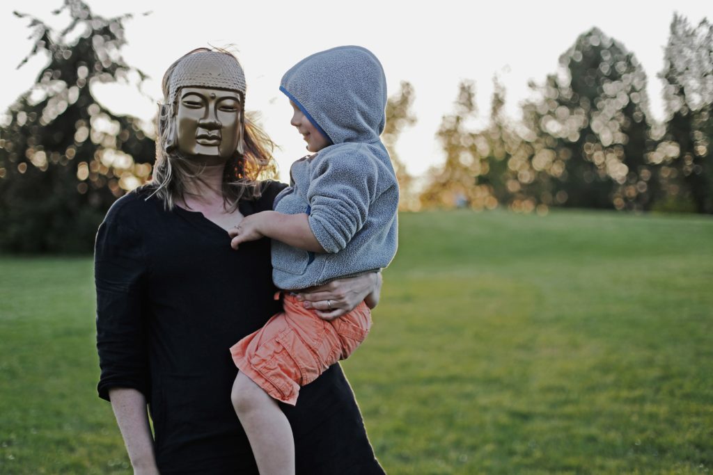 A mother with blond hair holds a small child. She wears a golden Buddha mask.