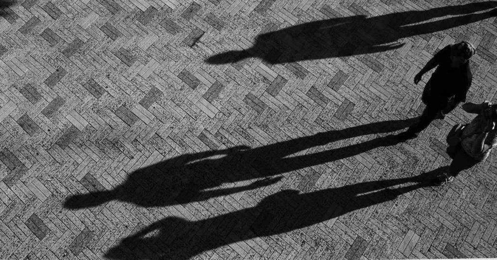 Two men are shown walking on a concrete floor, with three large shadows, indicating the incompleteness of the provisional life.