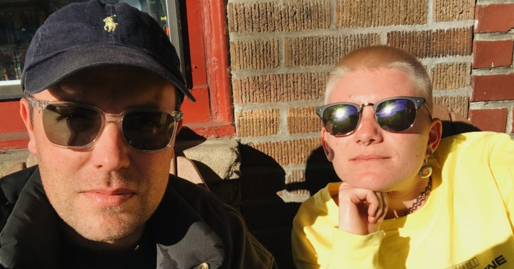 A photograph of No Small Thing podcast hosts Scott Gronholz and Marie. Both wear sunglasses and are shown against a brick wall.