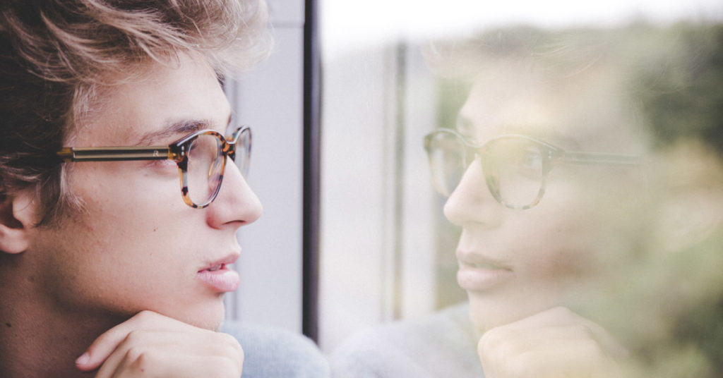 a blond man wearing tortoiseshell glasses stares out the window. His reflection stares back, illustrating the topic of self-reflection.
