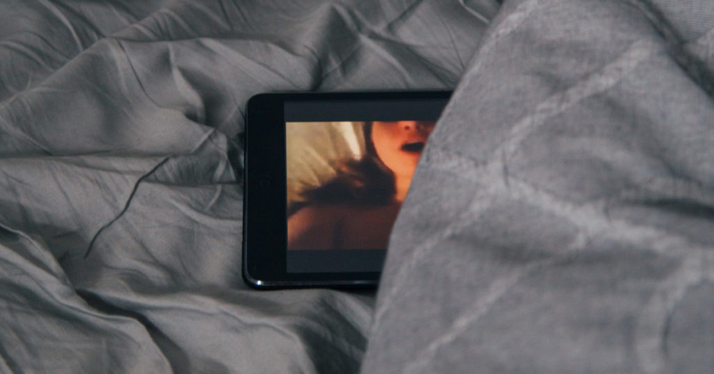 A laptop sits on some grey sheets, showing an image of a breathless woman on the screen, illustrating the topic of pornography.