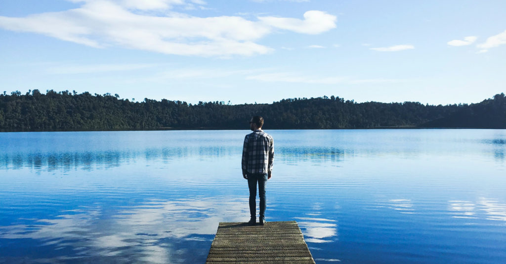 A man stands on a small pier over a lake, considering the nature of reality.